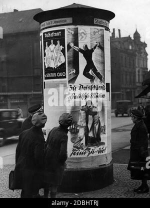 "An einer der Werbesäulen an der Ecke St. Johannerstraße in Saarbrücken schauen Passanten auf Wahlplakate für das Referendum über die Angliederung der Saar an das Deutsche Reich am 13.01.1935. Links das Wahlplakat der separatistischen Volksfront. Drei Männer, die bereit sind, sich zurückzuschlagen, stehen Arm in Arm auf einer Karte des Saargebiets gegen das Dritte Reich. Bildunterschrift: ''Für Status Quo''. Rechts die Plakate der Deutschen Front. Oben: ''Nach Deutschland''. Ein Mann öffnet das Tor zum Dritten Reich, symbolisiert durch das Hakenkreuz. Unten: ''Deutsche Mutter. Home to You!''' Stockfoto