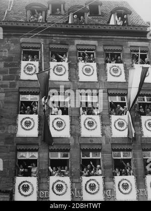 Blick auf die Außenfassade eines mit dem Nürnberger Wappen und Nationalflaggen geschmückten Hauses auf dem so genannten Adolf-Hitler-Platz während des NSDAP-Kongresses in Nürnberg. Die Bewohner des Gebäudes stehen in den Fenstern und führen den Hitlergruß vor. Links ist eine Tafel angebracht, die darauf hinweist, dass Willibald Pirckheimer, ein Nürnberger Humanist des späten Mittelalters, in diesem Gebäude einst lebte. Stockfoto