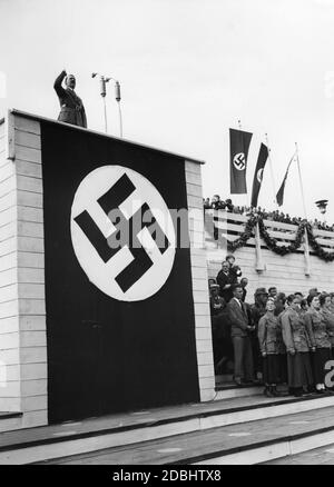 Adolf Hitler hält während der Parade des NS-Arbeitsdienstes eine Rede vom mit Hakenkreuzen geschmückten Rednerstand auf dem Zeppelinfeld auf dem Reichsparteitagsgelände. Rechts von der Hakenkreuzflagge hängt die Flagge des Rad. Unter der Tribüne befinden sich mehrere Frauen des Arbeitsdienstes. Stockfoto