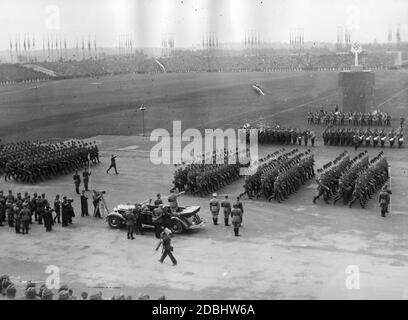 Adolf Hitler grüßt in seinem Mercedes die Formationen des Reichsarbeitsdienstes auf dem Zeppelin-Feld. Links von ihm filmt ein Kameramann die Szene. Im Hintergrund befindet sich das Denkmal der Arbeit. Davor gibt es eine Blaskapelle und eine Gruppe von Trommlern. Stockfoto