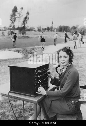 Eine Frau mit Mikrofon arbeitet an einer Telefonzentrale. Vermutlich wechselt sie Anrufe, indem sie die Schalter auf der Box umdreht. Im Hintergrund wird auf dem Golfplatz Golf gespielt. Stockfoto