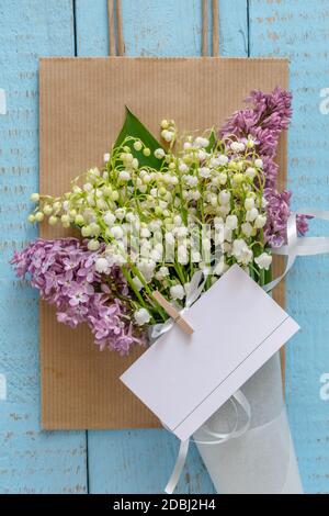 Maiglöckchen blüht mit Flieder in einer Papiertüte auf blauem Hintergrund. Flach liegend, Draufsicht, Kopierbereich Stockfoto