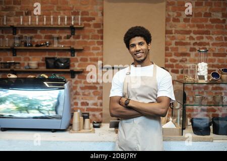 Lächelnder junger afroamerikanischer Kerl im Schürze, mit gekreuzten Händen in der Nähe der Theke mit Kaffeeausrüstung Stockfoto