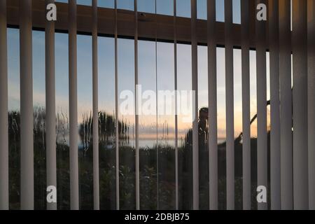 Vertikale Lamellenrollos an einem Fenster von innen gesehen, während ein Fensterputzer reines Wasser verwendet, um das Fenster zu reinigen. Stockfoto