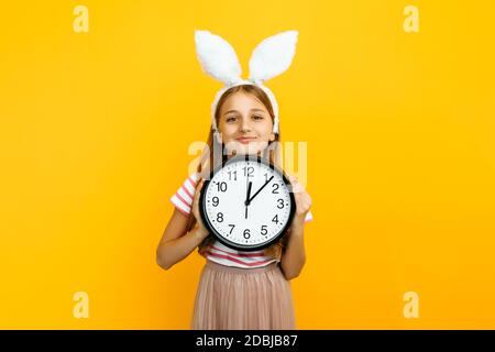 Happy schönes Mädchen auf dem Kopf mit Kaninchenohren, mit einer Wanduhr in den Händen auf einem gelben Hintergrund. Symbol von Ostern und Frühling. Stockfoto