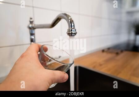 mans Hand hält ein leeres Glas in der Nähe einer Edelstahl-Küchenarmatur. Person erwägen Ausgießen von fließendem Wasser aus Chrom Wasserhahn. Trinkbar oder nicht-potabl Stockfoto