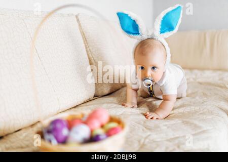 Niedliches lustiges Baby mit Kaninchenohren kriecht in den Korb Mit Ostereiern zu Hause auf dem Sofa Stockfoto