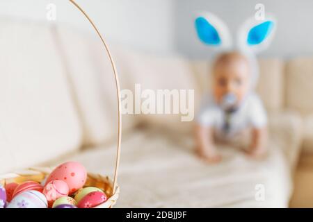 Niedliches lustiges Baby mit Kaninchenohren kriecht in den Korb Mit Ostereiern zu Hause auf dem Sofa Stockfoto