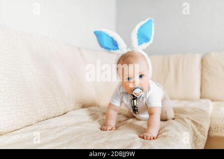 Niedliches lustiges Baby mit Kaninchenohren kriecht in den Korb Mit Ostereiern zu Hause auf dem Sofa Stockfoto