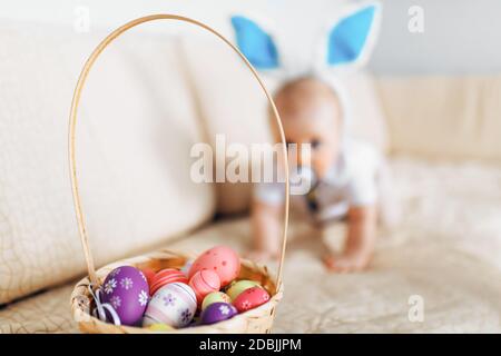 Niedliches lustiges Baby mit Kaninchenohren kriecht in den Korb Mit Ostereiern zu Hause auf dem Sofa Stockfoto
