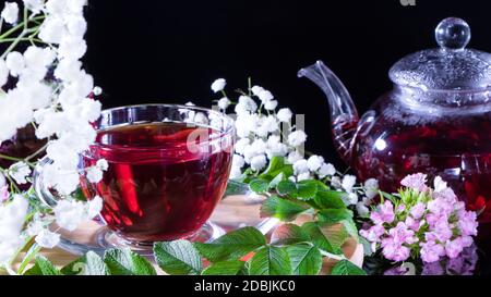 Ein Becher roten Tees und eine Teekanne in weißen Hibiskusblüten und grünen Blättern medizinischen Tees auf einem Holzständer.Zen-Tee Zeremonie. Foto von roten Kräuter Indi Stockfoto