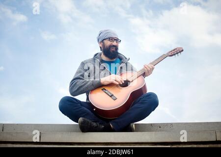 Der fröhliche bärtige Mann spielt Gitarre und sitzt auf der Treppe im Freien auf bewölktem Himmel Hintergrund. Stockfoto