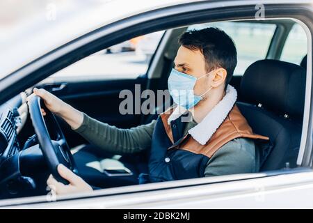 Schöner junger maskierter Mann im Auto sitzend, Schutzmaske gegen Coronavirus, Fahrer auf einer Stadtstraße während eines Coronavirus-Ausbruchs, covid-19 Stockfoto