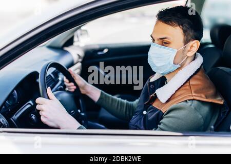 Schöner junger maskierter Mann im Auto sitzend, Schutzmaske gegen Coronavirus, Fahrer auf einer Stadtstraße während eines Coronavirus-Ausbruchs, covid-19 Stockfoto