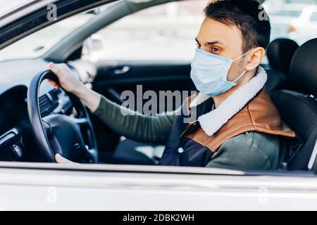 Schöner junger maskierter Mann im Auto sitzend, Schutzmaske gegen Coronavirus, Fahrer auf einer Stadtstraße während eines Coronavirus-Ausbruchs, covid-19 Stockfoto