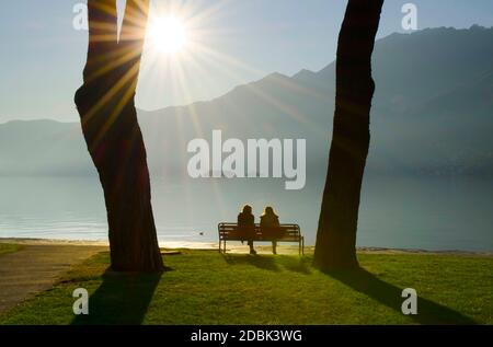 Ehepaar sitzt auf einer Bank am Ufer des Lago Maggiore mit Brissago Inseln und Berg mit Sunbeam in Ascona, Tessin in der Schweiz. Stockfoto