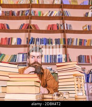 Lehrer oder Schüler mit Bart sitzen am Tisch mit Sanduhr und Brille, defokussed. Bookworm-Konzept. Mann auf strengem Gesicht sitzen zwischen Haufen von Büchern, während das Studium in der Bibliothek, Bücherregale auf Hintergrund. Stockfoto