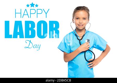 Kind im Kostüm des Arztes hält Stethoskop während des Stehens mit Hand auf Hüfte fast Happy Labor Day Schriftzug auf weiß Stockfoto