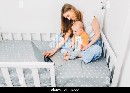 Eine junge Mutter im Schlafanzug mit einem kleinen Kind, sitzt auf dem Bett, mit einem tragbaren Laptop. Das Konzept der Remote-Arbeit in Quarantäne Stockfoto