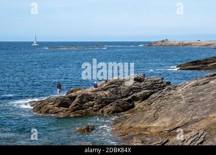 Mann, der auf den Felsen mit einer weiblichen Begleitung und einer vorbeifahrenden Yacht angeln kann, bei der Cote Sauvage auf der Halbinsel Quiberon, Bretagne, Frankreich. Stockfoto