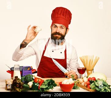 Kochen mit Lächeln in burgunderroten Uniform sitzt am Küchentisch mit Gemüse und Geschirr. Konzept der Restaurantküche. Der Küchenchef bereitet das Essen zu. Mann mit Bart hält Pilz und Messer auf weißem Hintergrund Stockfoto