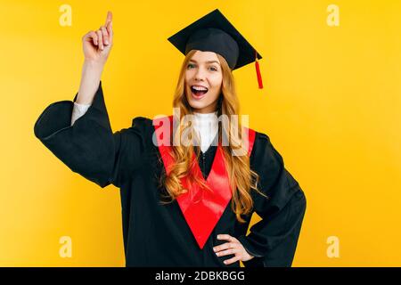 Eine attraktive Absolventin hat eine tolle Idee, hebt ihren Zeigefinger in der Eureka-Geste, hält den Atem an und öffnet ihren Mund auf gelbem Hintergrund Stockfoto