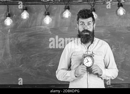 Lehrer in Brillen hält Wecker. Zeitplan und Regime Konzept. Mann mit Bart und Schnurrbart auf überrascht Gesicht im Klassenzimmer. Bärtige hipster hält, schwarzen Brettes auf Hintergrund, kopieren. Stockfoto