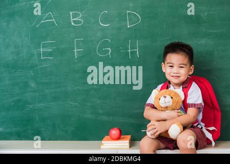 Zurück zur Schule. Glücklich asiatisch lustig niedlich kleinen Kind junge aus dem Kindergarten in Schüler Uniform mit Schultasche lächelnd und umarmend Teddybär auf grün s Stockfoto