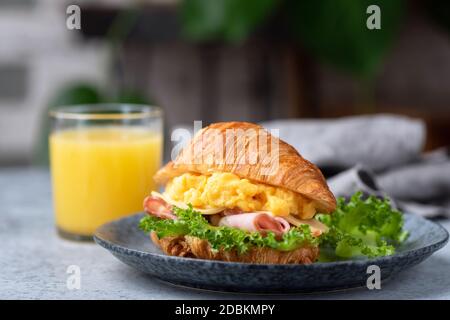 Köstliches Croissant-Sandwich mit Schinken, Käse und Rührei auf dem Teller serviert mit einem Glas Orangensaft Stockfoto