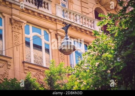 Nahaufnahme einer Straßenlaterne auf Luftdrähten über grünen Bäumen und einem schönen alten Gebäude im Hintergrund. Hängende Straßenbeleuchtung in Budapest City Cent Stockfoto