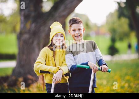 Kaukasische Kinder Zwillinge Junge und Mädchen Schulkinder alt zusammen bei sonnigem Wetter im Herbst Park nach dem Reiten auf Kick Scooter. Glückliche Kinder ridin Stockfoto