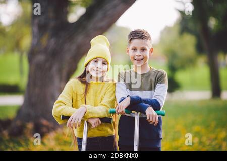 Zwei Kinder kaukasischen Jungen und Mädchen Zwillinge posieren mit Motorroller im Herbst Park. Glückliche Kinder reiten Kick Scooter im öffentlichen Park. Kindersport. Outdoor-Act Stockfoto