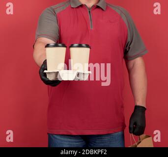 Mann in einem roten T-Shirt, trägt schwarze Latexhandschuhe, hält ein Tablett mit Einweg-Tassen Kaffee und eine Papiertasche aus braunem Kraftpapier, roter Hintergrund Stockfoto