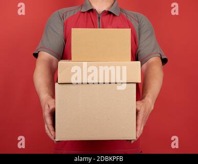 Erwachsene Mann in einem T-Shirt hält einen Stapel von großen Karton braunen Boxen auf einem roten Hintergrund, Konzept der Lieferung von Waren, bewegen Stockfoto