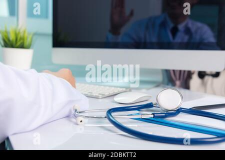 Mediziner arbeitet am Schreibtisch. Nahaufnahme des Stethoskops. Hand des Mannes Arzt Videoanruf Heben Sie die Hände, um Patienten auf Tisch Front Computer moni begrüßen Stockfoto