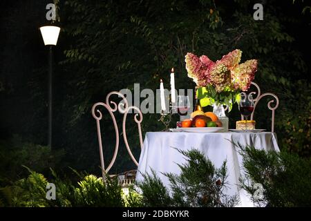 Ein Tisch mit weißer Tischdecke ist wunderschön im Nachtgarten eingerichtet. Hortensien auf einem Tisch in einer Vase.das Konzept eines romantischen Abends. Liebe und VA Stockfoto