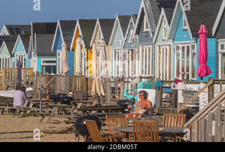 Menschen sitzen in der Sonne Sonnenbaden vor bunten Strandhütten auf Mudeford Sand Spit, Christchurch Großbritannien Stockfoto