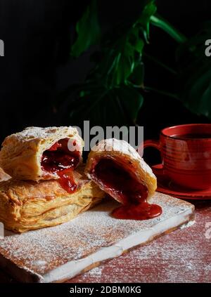 Fresh Puff mit Pflaume oder rote Johannisbeere Marmelade auf dem Tisch mit einer roten Tasse und Glas Marmelade auf dem schwarzen Hintergrund besetzt. Stockfoto