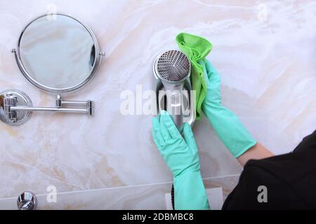 Eine Frau in einem Reinigungsservice Uniform wischt einen Haartrockner im Badezimmer. Nicht erkennbares Foto. Das Konzept der Sauberkeit und Hygiene. Speicherplatz kopieren. Stockfoto