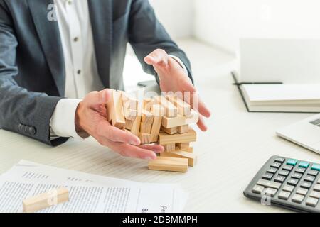 Beschnittene Ansicht des Geschäftsmannes hält Blöcke Holz Spiel zusammenbrechen, während in der Nähe von Dokumenten am Arbeitsplatz sitzen Stockfoto