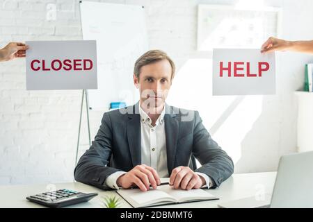 Geschäftsmann, der die Kamera anschaut, während er am Arbeitsplatz in der Nähe von Schildern mit geschlossenen und Hilfsbeschriftungen auf verschwommenem Hintergrund sitzt Stockfoto