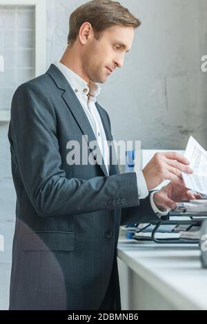 Seitenansicht eines fokussierten Geschäftsmannes, der durch die Papiere schaut, während er in der Nähe des Dokumententablageblätes auf der Fensterbank steht Stockfoto