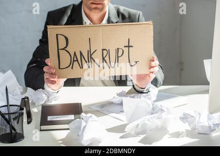 Beschnittene Ansicht eines Geschäftsmannes, der Karton mit bankrotten Schriftzügen hält, während er mit zerknittertem Papier am Schreibtisch sitzt Stockfoto