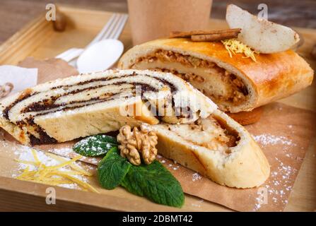 Mohn- und Apfelstrudel auf einem rustikalen Holztablett. Das Dekor der Duftrollen besteht aus Zimt, Minze, Nuss, Zitronenschale und Puderzucker. Nahaufnahme, verschwommen Stockfoto