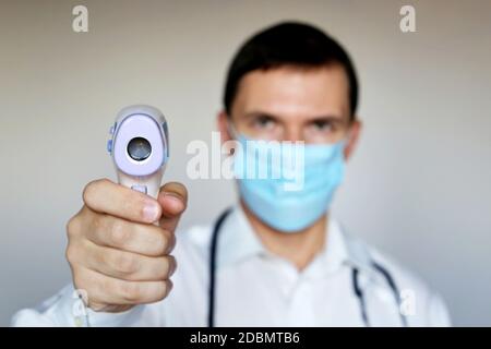 Mann in medizinischer Maske misst Körpertemperatur, Coronavirus-Symptome. Arzt mit einem digitalen isometrischen berührungslosen Thermometer in der Hand Stockfoto