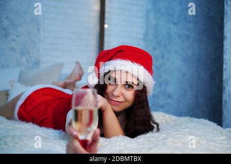 Junge Frau im Weihnachtsmann Kostüm mit einem Glas Champagner auf dem Bett liegend. Charmante weibliche Ruhe und Weihnachten zu Hause zu feiern. Konzept des Urlaubs Stockfoto