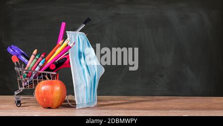 Zurück zur Schule während der Coronavirus-Pandemie mit Bleistiften, Buntstiften und Gesichtsmaske im Warenkorb mit Apfel gegen Tafel Stockfoto