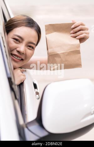 Asiatische junge Erwachsene im Auto hält Einweg-Tasche für nehmen aus Essen aus Drive-Thru Service-Restaurant. Drive Thru ist neu normal und beliebt Service hinten Stockfoto