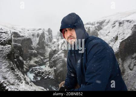 Ein junger Mann, der eine blaue Jacke trägt, kniet sich während eines Schneesturms zu einem Portrait in der Schlucht Fjaðrárgljúfur nieder. Stockfoto
