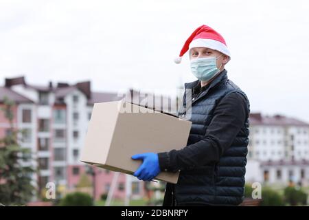 Delivery Mann in Schutzmaske, Handschuhe und santa Hut halten Box in den Händen im Freien, Lieferservice während Coronavirus in der Ferienzeit Stockfoto